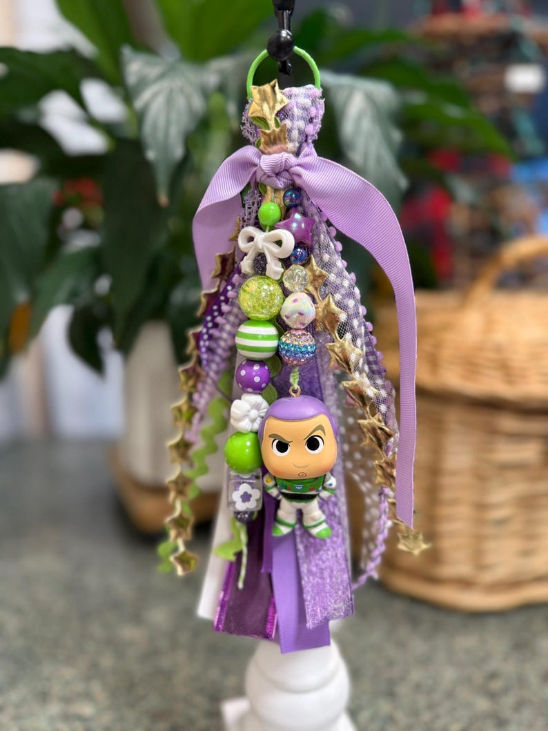 May include: A decorative keychain featuring a Buzz Lightyear figurine. The keychain is adorned with purple ribbons, beads, and gold star accents. A green loop and black clasp are at the top.