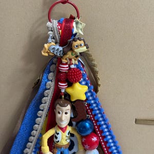Op de afbeelding: Een decoratieve sleutelhanger met een rode karabijnhaak en ring, versierd met gelaagde linten in blauw, rood en wit. Het bevat een plastic Woody-figuur, een bruine hondfiguur, een gele ster-kraal en diverse rode, blauwe en witte ronde kralen.