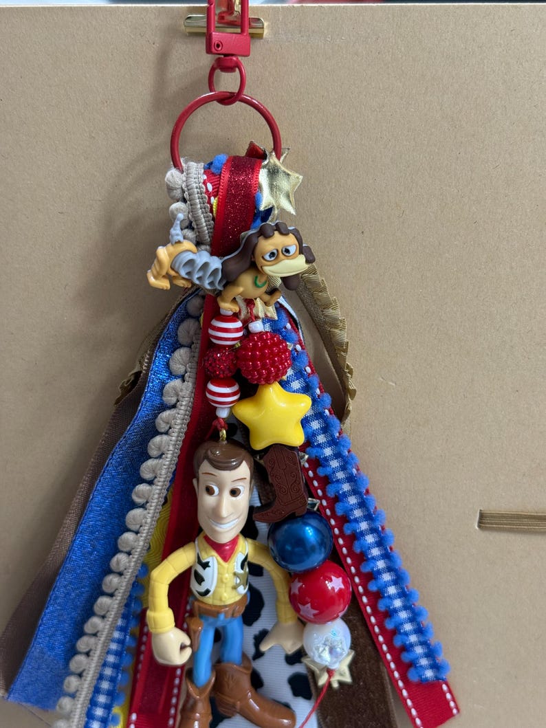 Op de afbeelding: Een decoratieve sleutelhanger met een rode karabijnhaak en ring, versierd met gelaagde linten in blauw, rood en wit. Het bevat een plastic Woody-figuur, een bruine hondfiguur, een gele ster-kraal en diverse rode, blauwe en witte ronde kralen.