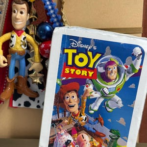 Op de afbeelding: Een Disney Toy Story VHS-hoes met Woody en Buzz Lightyear en andere personages, naast een hangend Woody-figuurtje met een gele ster, blauwe en rode kralen en een bruine cowboylaars-bedel op een achtergrond van linten.