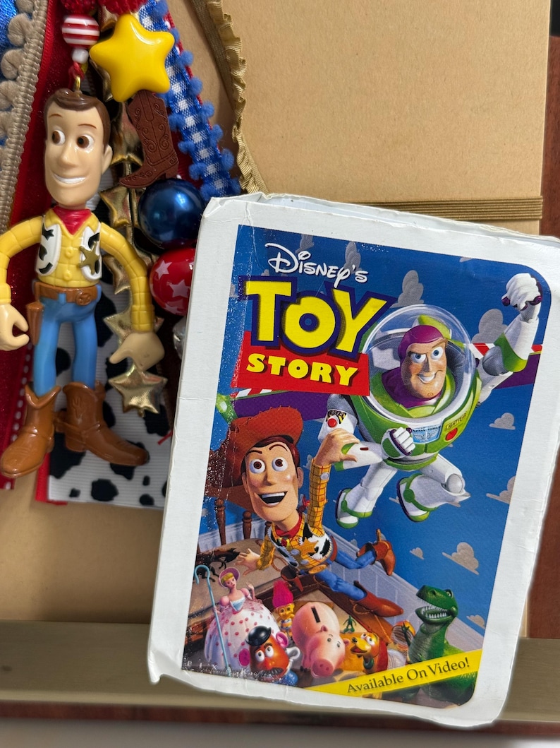 Op de afbeelding: Een Disney Toy Story VHS-hoes met Woody en Buzz Lightyear en andere personages, naast een hangend Woody-figuurtje met een gele ster, blauwe en rode kralen en een bruine cowboylaars-bedel op een achtergrond van linten.