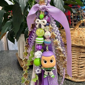 May include: A handmade purple and green keychain featuring a Buzz Lightyear figurine. The keychain includes a variety of beads, ribbons, and star-shaped accents. The design incorporates shades of purple, green, white, and gold.