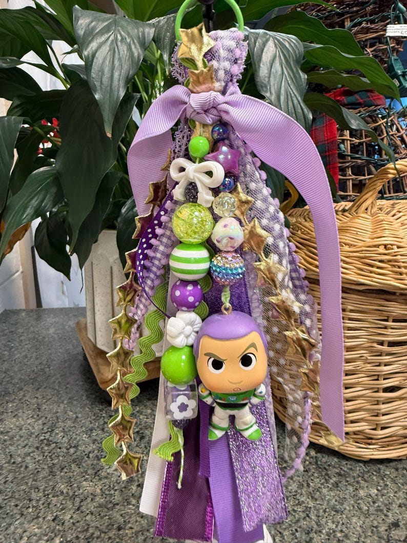 May include: A handmade purple and green keychain featuring a Buzz Lightyear figurine. The keychain includes a variety of beads, ribbons, and star-shaped accents. The design incorporates shades of purple, green, white, and gold.
