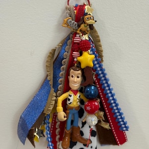 Op de afbeelding: Een decoratieve hangende versiering met een Woody-figuurtje en een Slinky Dog-bedel bevestigd aan een bundel veelkleurige linten. De linten bevatten blauwe glitter, rode, witte en zwart-witte koeienprintpatronen, geaccentueerd met stervormige bedels en kralen.