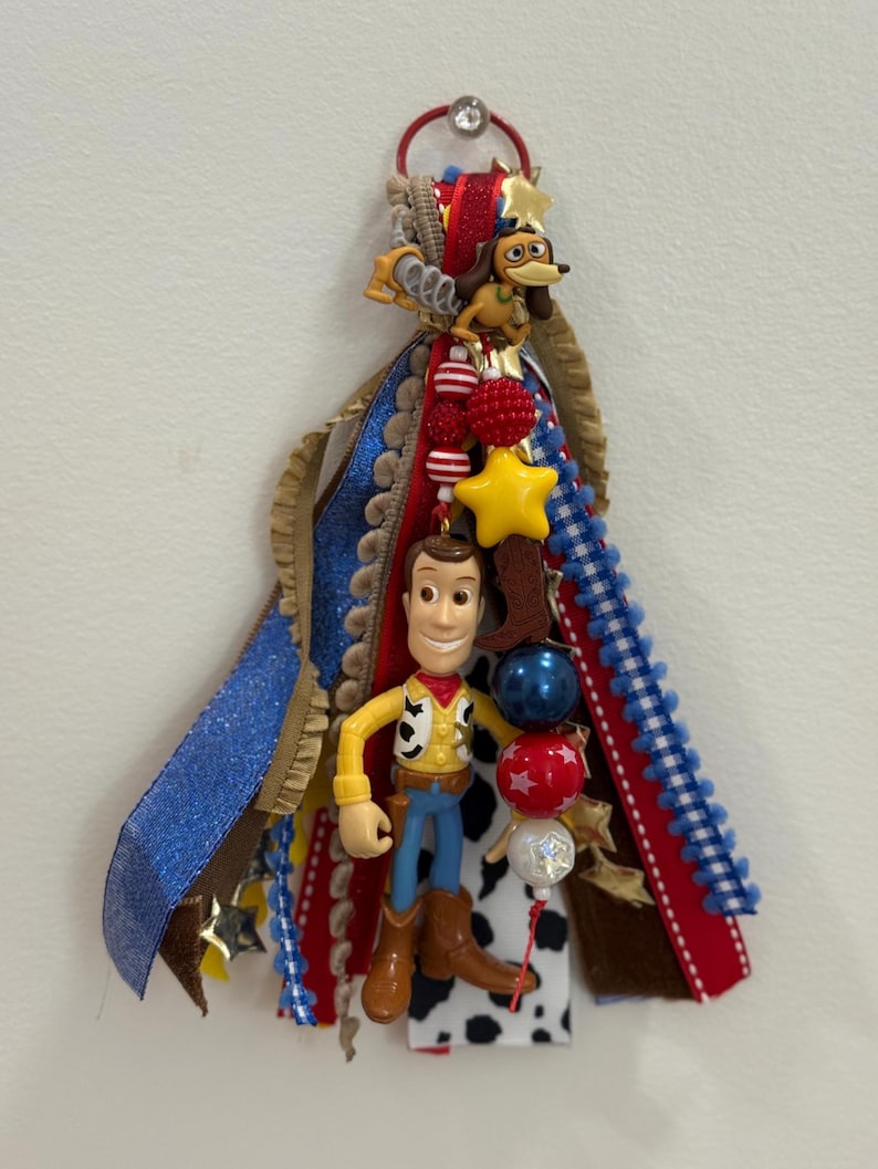 Op de afbeelding: Een decoratieve hangende versiering met een Woody-figuurtje en een Slinky Dog-bedel bevestigd aan een bundel veelkleurige linten. De linten bevatten blauwe glitter, rode, witte en zwart-witte koeienprintpatronen, geaccentueerd met stervormige bedels en kralen.