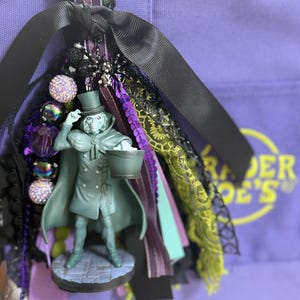 May include: A decorative bag charm featuring a miniature figure of a ghost in a top hat and cape holding a bucket. The charm is adorned with black, purple, and green ribbons, sequined fabric, lace, and various textured beads attached to a metal ring.