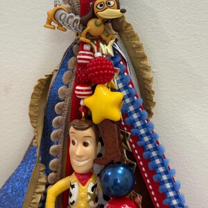 Op de afbeelding: Een decoratief homecoming mum-accessoire met een plastic Woody-figuurtje, een Slinky Dog-bedel en een cowboylaars-bedel. Het stuk is versierd met rode, witte, blauwe en gouden linten, samen met diverse kralen, sterren en pompon-afwerkingen.