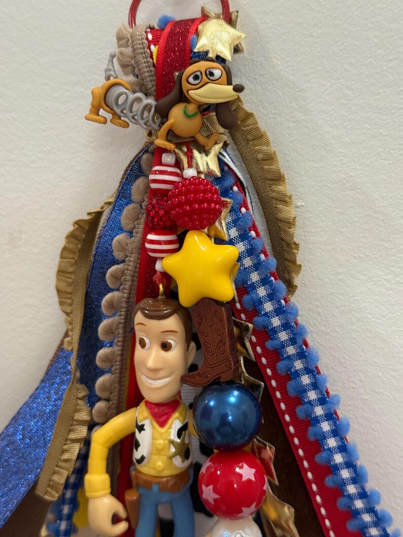 Op de afbeelding: Een decoratief homecoming mum-accessoire met een plastic Woody-figuurtje, een Slinky Dog-bedel en een cowboylaars-bedel. Het stuk is versierd met rode, witte, blauwe en gouden linten, samen met diverse kralen, sterren en pompon-afwerkingen.