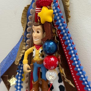 Op de afbeelding: Een decoratieve hangende versiering met een plastic Woody-figuurtje uit Toy Story, omringd door gelaagde linten in blauw, rood, wit en koeienprint. Het ornament bevat een gele ster, een bruine laarsbedel en rode, blauwe en witte kralen.