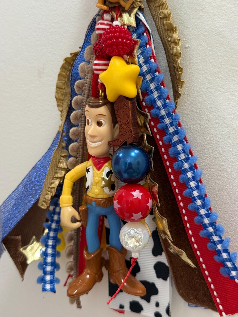 Op de afbeelding: Een decoratieve hangende versiering met een plastic Woody-figuurtje uit Toy Story, omringd door gelaagde linten in blauw, rood, wit en koeienprint. Het ornament bevat een gele ster, een bruine laarsbedel en rode, blauwe en witte kralen.