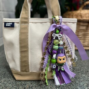May include: A small canvas tote bag with tan accents and a decorative charm. The charm features a Buzz Lightyear figurine, beads, ribbons, and star-shaped embellishments in shades of purple, green, and gold.