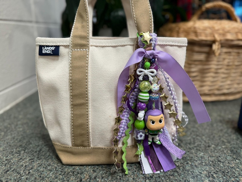 May include: A small canvas tote bag with tan accents and a decorative charm. The charm features a Buzz Lightyear figurine, beads, ribbons, and star-shaped embellishments in shades of purple, green, and gold.