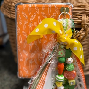 May include: An orange phone case with a white carrot pattern, adorned with a yellow polka dot ribbon and a beaded charm. The charm includes green, orange, and white beads, and a small flower.