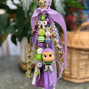 May include: A decorative tassel featuring a Buzz Lightyear figurine. The tassel includes purple ribbons, gold star accents, and various beads in shades of green, purple, and white. A green loop and a white base complete the design.