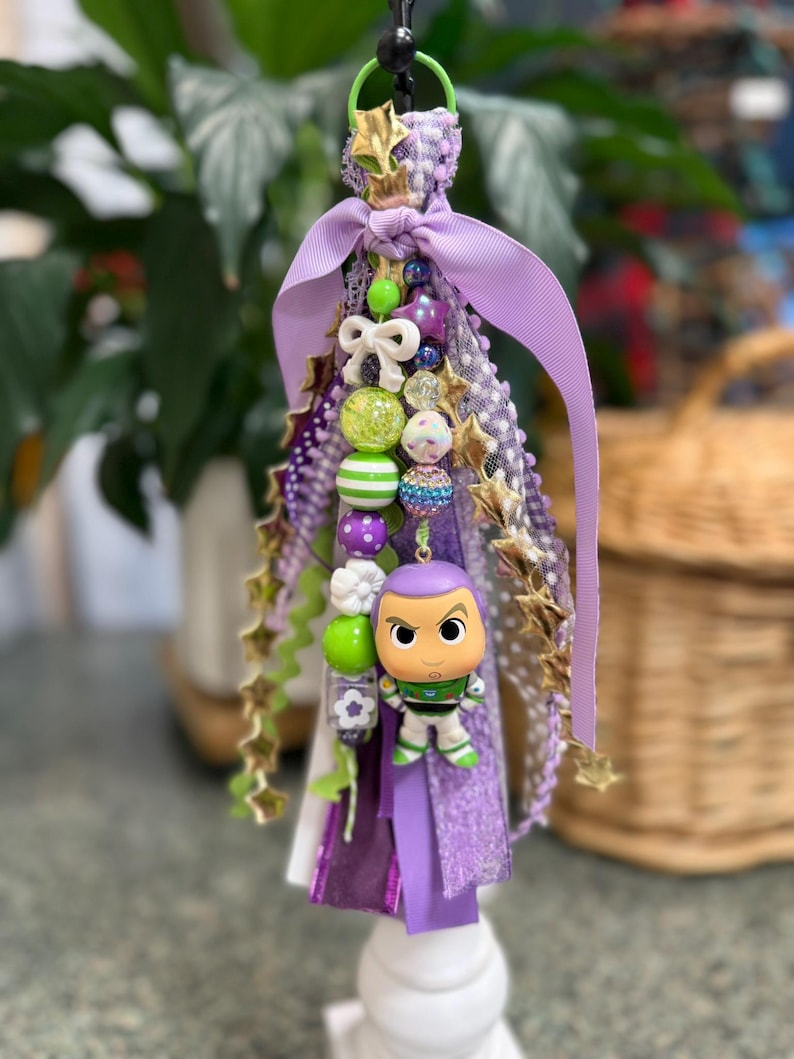 May include: A decorative tassel featuring a Buzz Lightyear figurine. The tassel includes purple ribbons, gold star accents, and various beads in shades of green, purple, and white. A green loop and a white base complete the design.