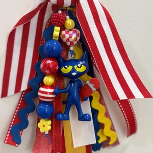 May include: A decorative tassel featuring a blue cat character holding a red skateboard, surrounded by multi-colored beads including a checkered heart, red stars, and yellow flowers, all attached to a cluster of red, white, blue, and yellow ribbons.