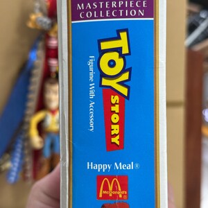Op de afbeelding: Een verticale rechthoekige doos voor een Toy Story Happy Meal figuurtje met accessoire uit de Walt Disney Masterpiece Collection. De blauwe verpakking bevat het Toy Story logo, het McDonald's logo en een illustratie van Woody.