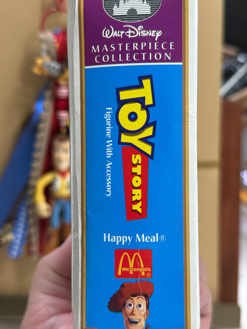 Op de afbeelding: Een verticale rechthoekige doos voor een Toy Story Happy Meal figuurtje met accessoire uit de Walt Disney Masterpiece Collection. De blauwe verpakking bevat het Toy Story logo, het McDonald's logo en een illustratie van Woody.