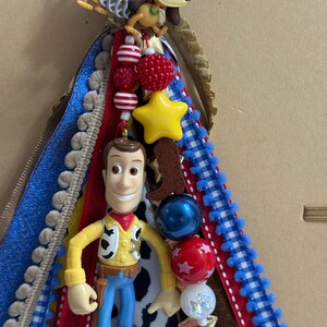 Op de afbeelding: Een decoratieve lintcluster met een plastic Woody-figuurtje en een Slinky Dog-bedel. Het arrangement bevat rode, blauwe en witte linten, een gele ster-kraal, een bruine cowboylaars-bedel en diverse kralen met patronen en texturen.