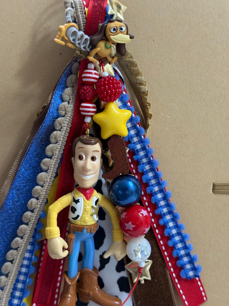 Op de afbeelding: Een decoratieve lintcluster met een plastic Woody-figuurtje en een Slinky Dog-bedel. Het arrangement bevat rode, blauwe en witte linten, een gele ster-kraal, een bruine cowboylaars-bedel en diverse kralen met patronen en texturen.