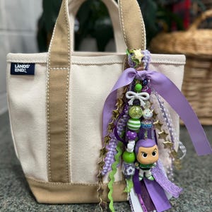 May include: A small canvas tote bag with tan accents and handles. The bag features a charm with purple, green, and gold accents, including a Buzz Lightyear figurine. The bag has the text "LANDS' END" on the front.