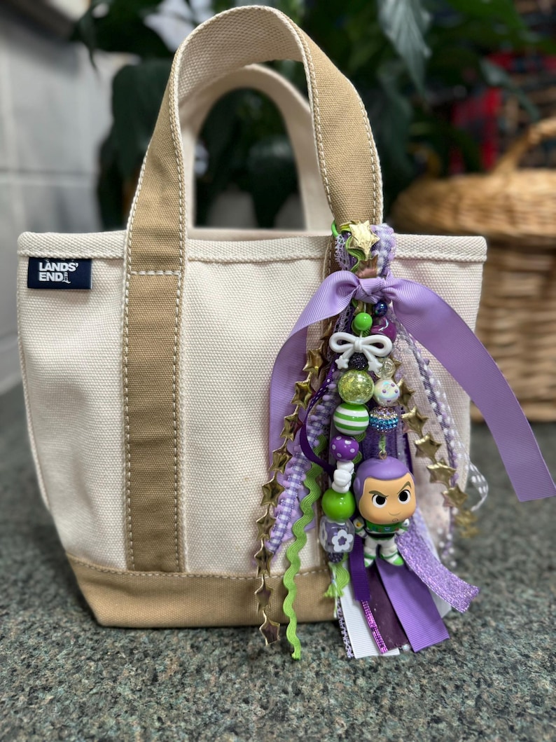 May include: A small canvas tote bag with tan accents and handles. The bag features a charm with purple, green, and gold accents, including a Buzz Lightyear figurine. The bag has the text "LANDS' END" on the front.