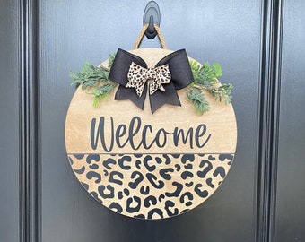 May include: A round wooden door hanger with a leopard print design and the word "Welcome" written in black. A black bow with a leopard print accent is attached to the top of the hanger.