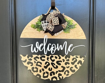 May include: A round wooden door hanger with a black and white leopard print design. The top half of the hanger is natural wood with a black ribbon bow and green leaves. The bottom half is black with the word "welcome" written in white.