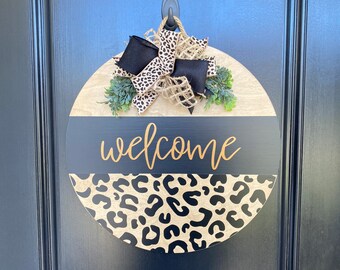 May include: A round wooden door hanger with a black and white leopard print design. The top half of the hanger is a natural wood color with a black bow and greenery. The bottom half is black with the word "welcome" written in gold lettering.