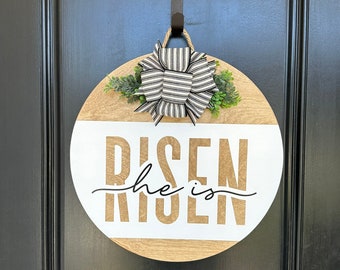 May include: A round wooden door hanger with a white background and the text "He is Risen" in gold lettering. The hanger is decorated with a black and white striped bow and green foliage.