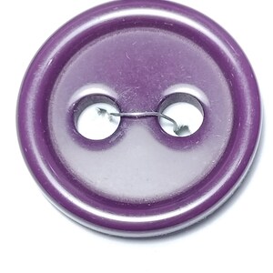 May include: Close-up of a round, purple button with two holes and a metal wire. The button has a glossy finish and is set against a white background. This sewing notion is suitable for various craft projects.