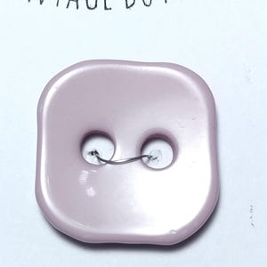 May include: A square, light pink vintage button with two holes and a wire loop. The button has a glossy finish and is set against a white background. The text "VINTAGE BUTTONS" is visible at the top.