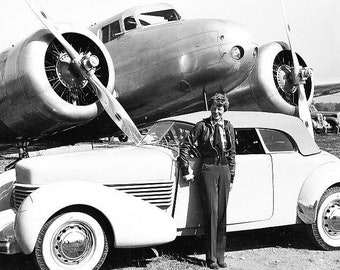 May include: Black and white photograph of a woman standing next to a cream-colored convertible car, with a large airplane in the background. The airplane has two propellers. The woman is wearing a leather jacket and pants.