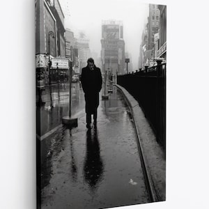 以下が含まれることがあります： 雨の降る街の道を歩く男性の白黒写真。男性はロングコートを着て、手にタバコを持っている。道は濡れていて、背景には建物が見える。