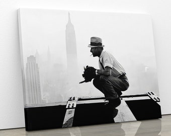May include: A black and white photograph of a man in a hat, sitting on a beam, looking out over a foggy cityscape. The man is holding a camera and appears to be taking a photograph.