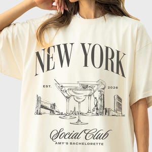 May include: An ivory short-sleeve t-shirt featuring a black graphic print. The design includes the text NEW YORK, Social Club, and AMY'S BACHELORETTE, along with illustrations of martini glasses and a city skyline with the text EST. 2026.