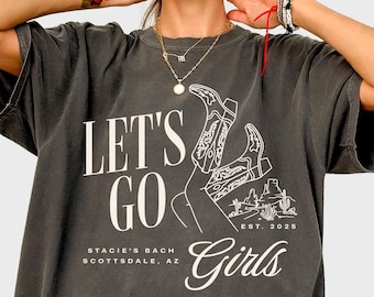 May include: A dark gray oversized t-shirt with a white graphic design that says "Let's Go Girls" with a drawing of a woman wearing cowboy boots. The text also says "Stacie's Bach Scottsdale, AZ Est. 2023"