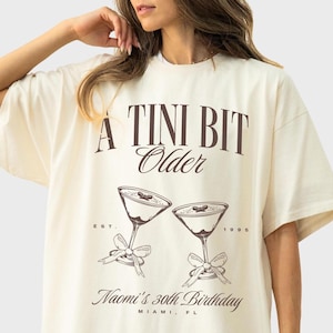 May include: A white t-shirt with a brown graphic that says "A Tini Bit Older" with two martini glasses and the text "Est. 1995 Naomi's 30th Birthday Miami, FL"