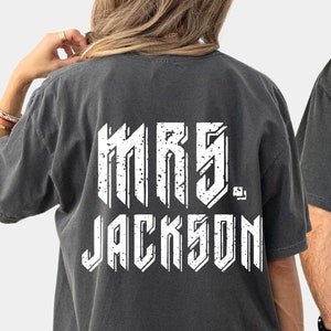May include: Two gray t-shirts with white text on the back. One shirt reads "MRS. JACKSON" and the other reads "MR. JACKSON". The shirts are a dark gray color and have a vintage, distressed look.