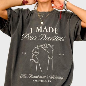 May include: Pepper-colored oversized t-shirt with the text "I Made Pour Decisions" in a script font. Below the text is an illustration of a wine bottle pouring into a glass. The shirt also includes the text "The Henderson's Wedding, Nashville, TN."