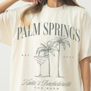 May include: Ivory oversized t-shirt with a graphic print of palm trees, a margarita glass, and the text "Palm Springs Est. 2024 Katie's Bachelorette The Bach"