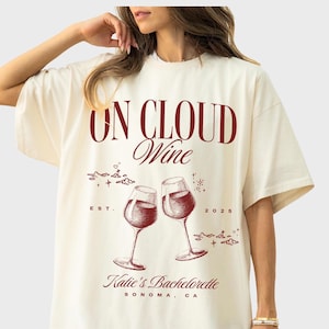 May include: A white oversized t-shirt with a red graphic design that says "On Cloud Wine" with two wine glasses clinking, "Est. 2025", and "Katie's Bachelorette Sonoma, CA".