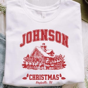 May include: White t-shirt with a red graphic featuring the word "JOHNSON" above a cabin scene with Santa and reindeer. The text "CHRISTMAS Nashville, TN" is below the cabin. The shirt is displayed on a white surface.
