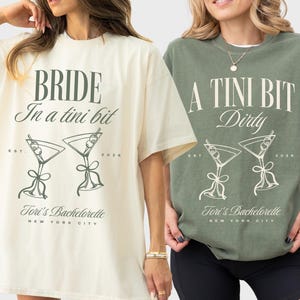 May include: Two oversized t-shirts, one ivory and one moss green, with graphic text and martini glass illustrations. The ivory shirt reads "BRIDE In a tini bit" and the green shirt says "A TINI BIT Dirty". Both shirts say "Tori's Bachelorette, New York City".