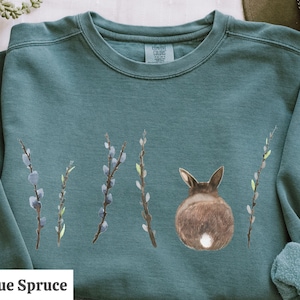 May include: A teal blue sweatshirt with a brown rabbit with a white tail, facing away from the viewer, and four sprigs of willow branches with green leaves and blue buds. The text "Blue Spruce" is printed in white at the bottom left of the sweatshirt.