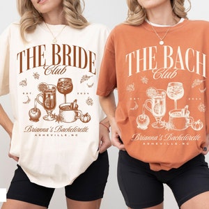 May include: Two women wearing t-shirts with a fall-themed design. The shirts say "The Bride Club" and "The Bach Club" with a graphic of drinks and fall foliage. The text below the graphic says "Brianna's Bachelorette Asheville, NC". The woman on the left is wearing a white shirt and the woman on the right is wearing a brown shirt.