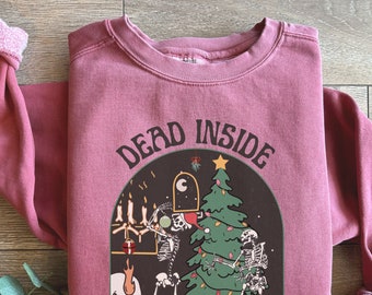 May include: A crimson sweatshirt with the text "Dead Inside" in black lettering. The design features a skeleton crew decorating a Christmas tree with ornaments and presents.