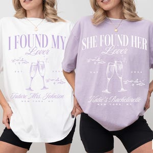 May include: Two white t-shirts with purple text and graphics. The left shirt says "I Found My Lover" with a graphic of two champagne glasses. The right shirt says "She Found Her Lover" with a graphic of two champagne glasses. The shirts are on models wearing black shorts. The left shirt is white and the right shirt is orchid.
