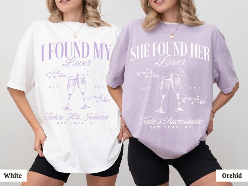 May include: Two white t-shirts with purple text and graphics. The left shirt says "I Found My Lover" with a graphic of two champagne glasses. The right shirt says "She Found Her Lover" with a graphic of two champagne glasses. The shirts are on models wearing black shorts. The left shirt is white and the right shirt is orchid.