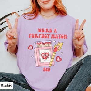 May include: A purple t-shirt with the text "We're a perfect match" and a graphic of a matchbox with a heart and a lit match. The text "Big" is below the graphic.